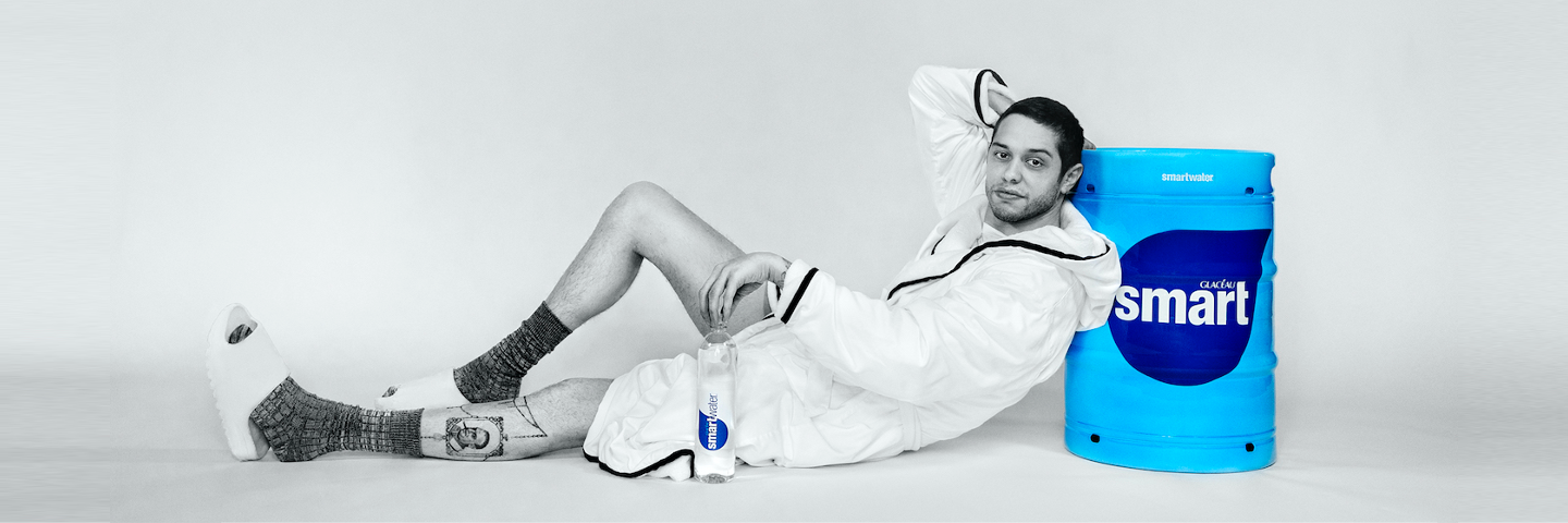 Comedian Pete Davidson leans on smartwater branded cooler while holding a bottle of smartwater