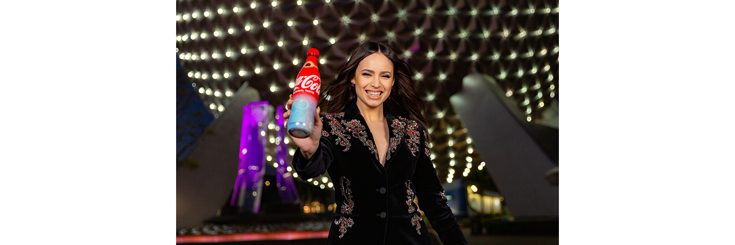 Sofia Carson stand in front of Epcot Center holding Walt Disney World 50th anniversary commerative Coca-Cola can. 