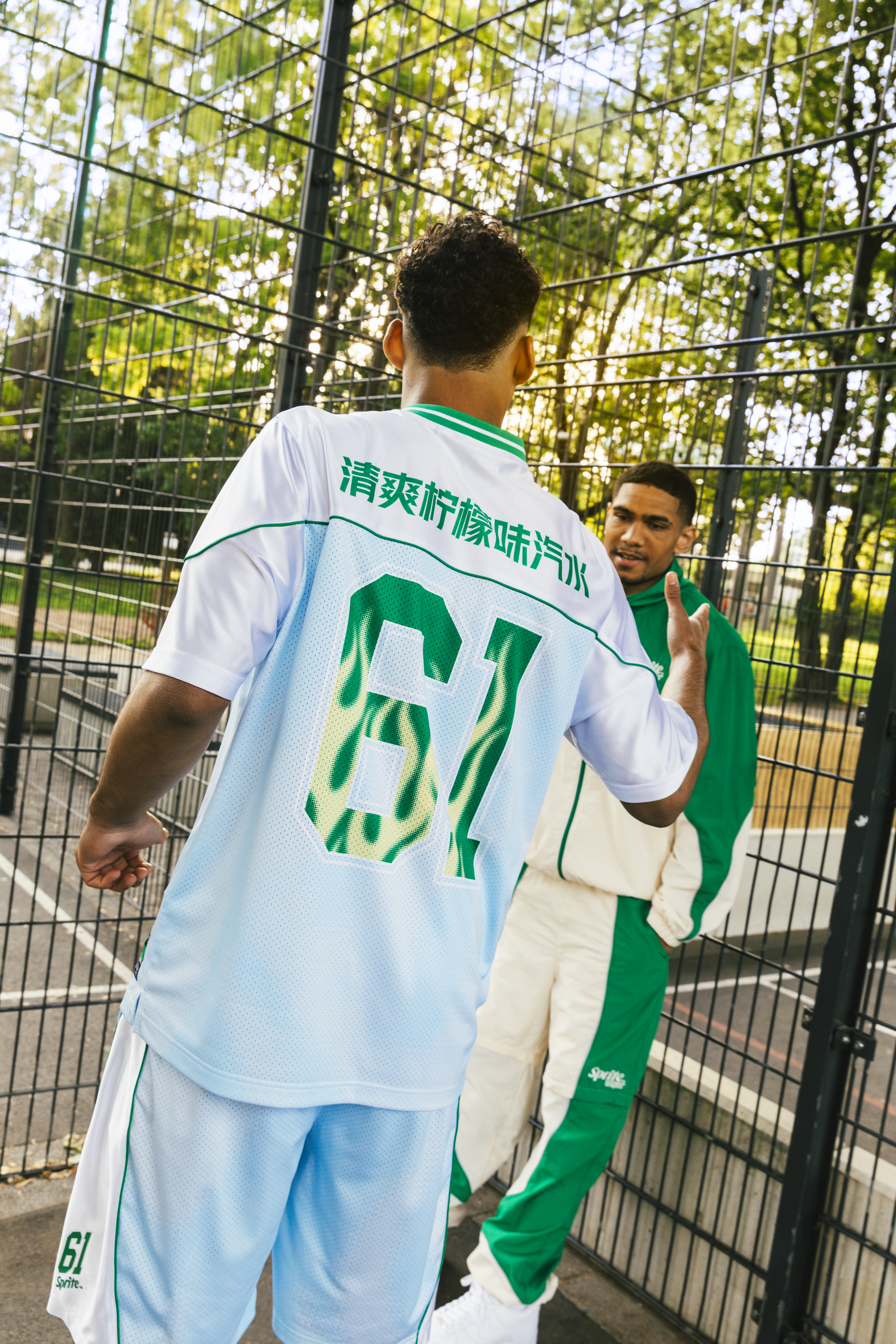 Two young men wearing SNIPES x Sprite gear hanging out by an outdoor basketball court fence