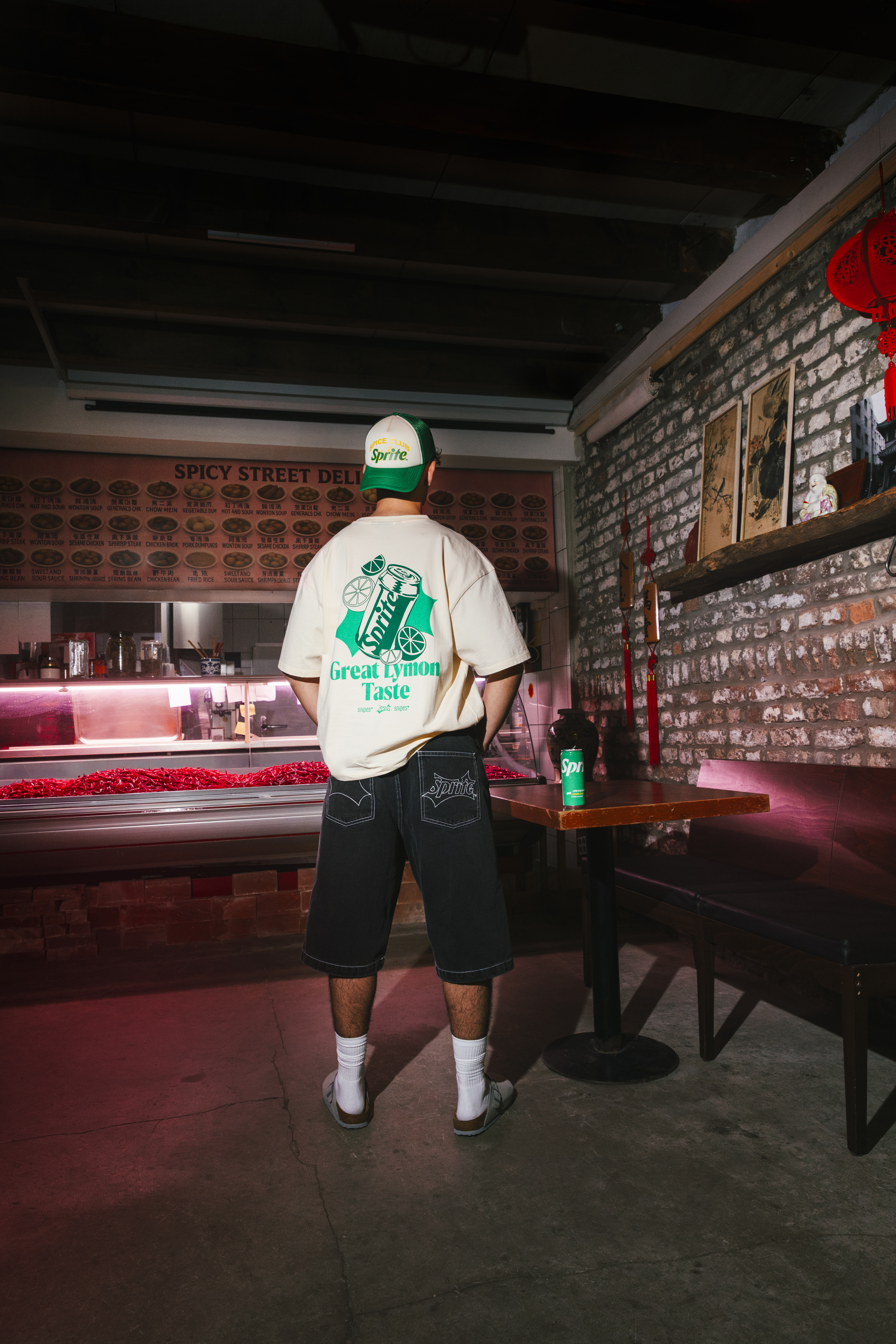 A man stands in a brick walled deli, back to camera, wearing SNIPES x Sprite shirt and denim shorts
