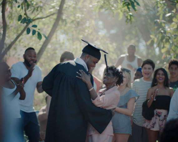 A man wearing graduation robe dances with an older woman as guests celebrate around themh