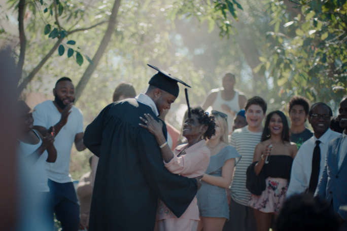 A man in graduation gown dancing with an older woman while surrounded by smiling guests