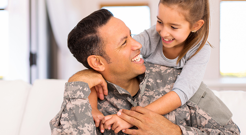 A man in military fatigues smiles at a young girl on his shoulders